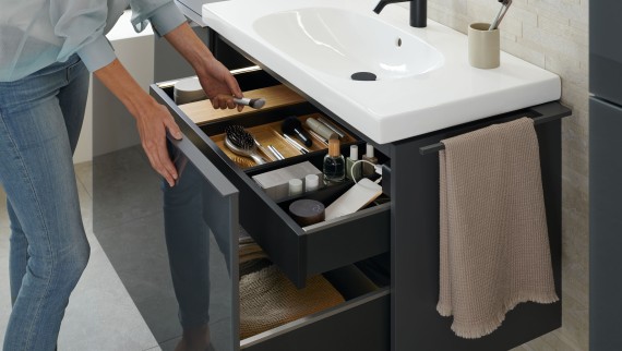 Une femme prend un pinceau dans un meuble lavabo Geberit Acanto ouvert en finition lave.