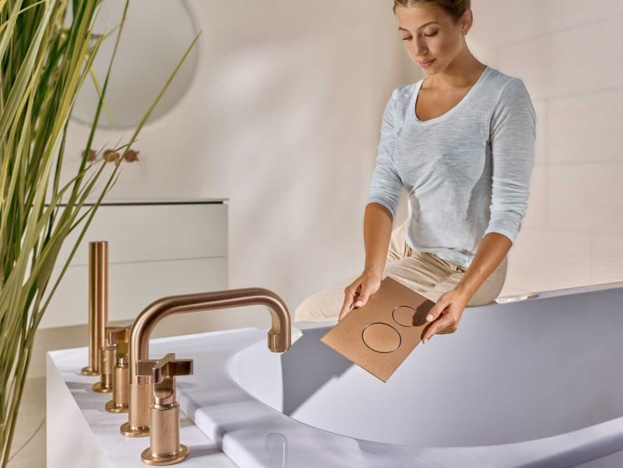 Femme dans la salle de bains avec une plaque de déclenchement Sigma40 en cuivre brossé, assortie à la robinetterie Femme dans la salle de bains avec une plaque de déclenchement Sigma40 en cuivre brossé, assortie à la robinetterie