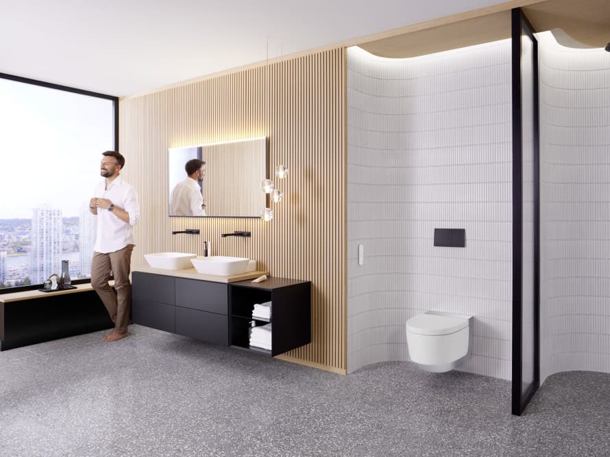 Homme dans une grande salle de bains avec des meubles sombres et des WC lavants Geberit AquaClean Homme dans une grande salle de bains avec des meubles sombres et des WC lavants Geberit AquaClean